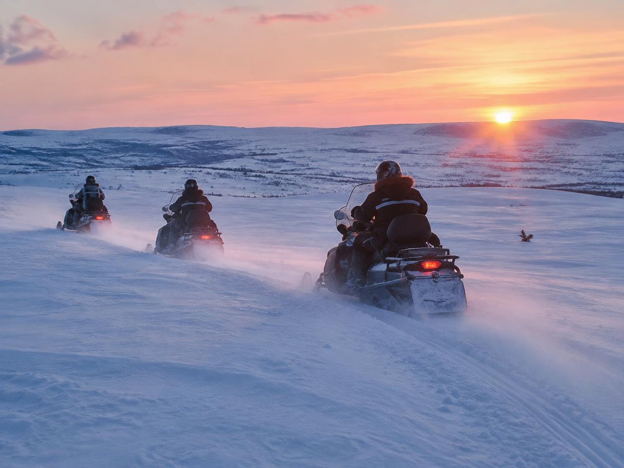 Tres personas conducen motos de nieve por una llanura nevada y abierta hacia un atardecer invernal con el sol bajo
