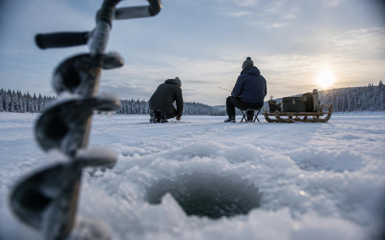 Una barrena para hielo en primer plano enmarca a dos personas que pescan en el hielo de un lago congelado