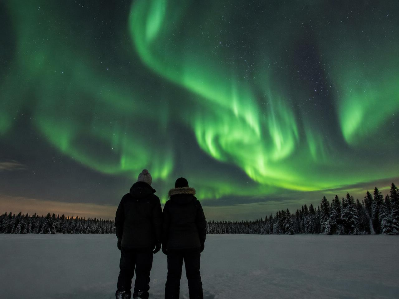 Dos personas a contraluz están de pie sobre la nieve contemplando unas auroras boreales de un verde brillante